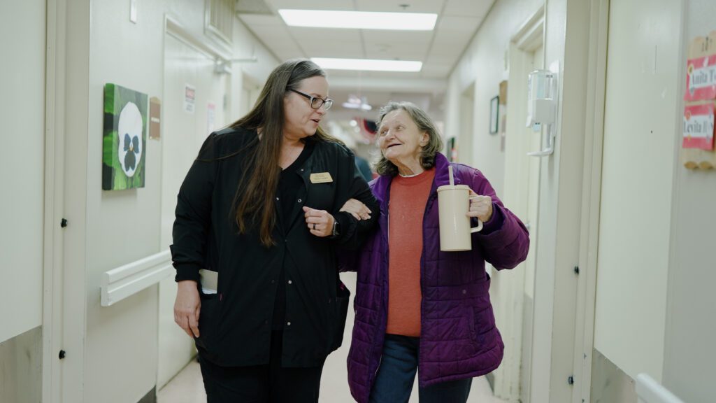 A woman smiles at a nurse assisting her down the hallway of a long-term care facility
