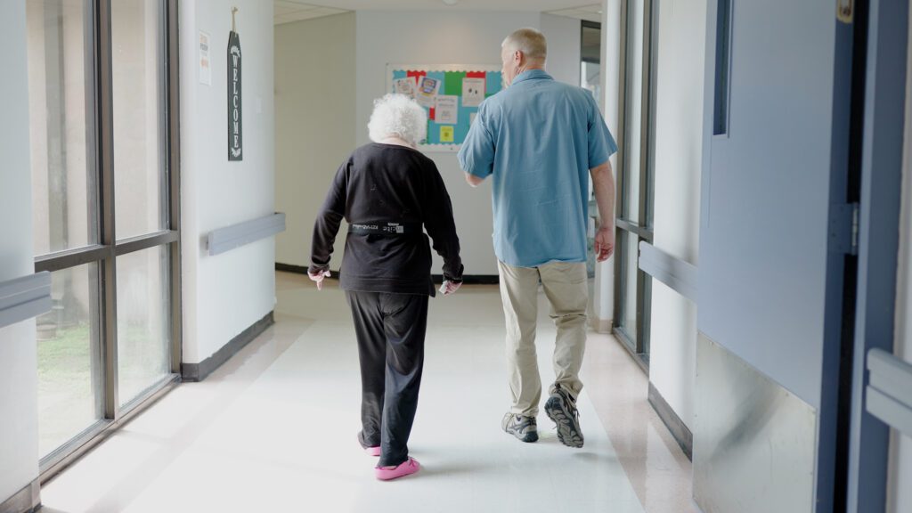 A physical therapist assisting a woman down the hallway of a skilled nursing facility in memory care